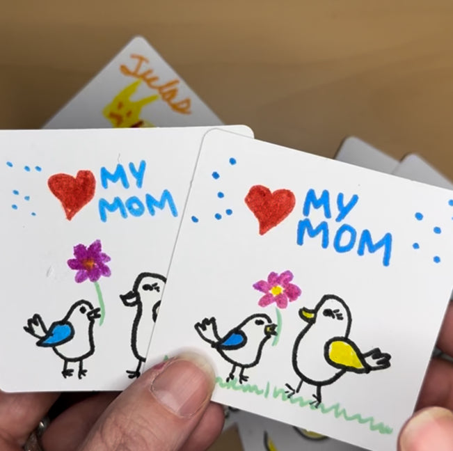 Hand holding a card with 'My Mom' and bird illustrations, with a blurred background.